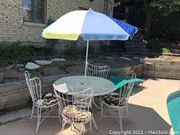 Full view of patio set with round white metal table, four matching metal chairs with floral cushions, large umbrella in blue, white, and green, and green cot beside the table.
