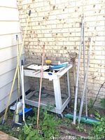 Multiple yard and pool tools standing against a wall, including brooms, a pool net, and a plastic rolling utility cart with shelves.