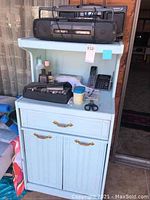 Painted wood baker's rack showing top shelves with Audiovox CD/radio/cassette player, RCA radio/cassette, cordless phone base, and various small items.