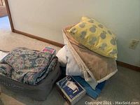 Stack of linens including patterned comforter in storage bag, decorative pillows, shearling throw, blankets and blue bedding items