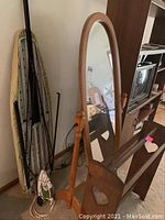 Oak adjustable oval floor mirror standing with wooden frame and supports, viewed angled to show tilt feature and surroundings including ironing board setup and ironing equipment nearby.