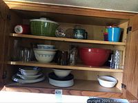 First photo of top cabinet shelves with glassware, dishware, plastic ware. Items include green plastic colander, glass mugs, bowls, plates, red plastic bowl, and storage containers.