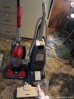 Photo showing a red Dirt Devil Power Max upright vacuum and a gray Hoover steam vac with hose and white Beam nozzle attachment laid on the carpeted floor.