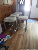 Four kitchen stools with chrome tubular metal frames and beige striped upholstered seats. One stool stained, shown stacked in line on hardwood floor near wooden doors.