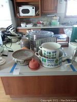 Kitchen countertop with various kitchen items including two white enameled pots with blue and green vegetable pattern, a ceramic bowl, terracotta garlic keeper, and other miscellaneous kitchenware items.