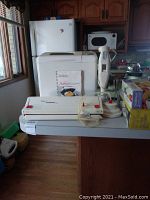 Wide shot showing all items on kitchen counter: Tilia Foodsaver vacuum sealer with replacement rolls, white Sunbeam bread maker with manual, and an immersion blender with stand.