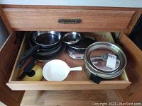 Top view of oak drawer containing six metal pots and pans with black handles, multiple glass lids, and a white CorningWare serving dish with handle.