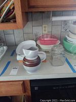 Kitchen counter displaying two white Corning baking dishes, ceramic pitcher with brown bands, glass jar, and pink plastic colander with Pyrex bowl underneath.