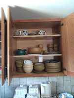 Open wood kitchen cabinet with shelves containing beige colored stoneware plates, bowls, patterned coffee mugs, shot glasses, and small teapot. Items arranged stacked and grouped.