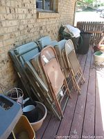 Group of seven folding chairs leaning against a brick wall on a wooden deck, showing both padded and metal types.