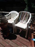 Photo of two white resin outdoor chairs stacked together, one with a colorful striped throw on the seat, situated on a wooden deck with plant pots around.