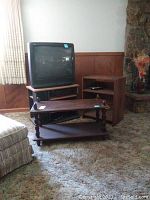 View of the 27-inch RCA analog CRT TV on the TV stand with two wooden corner shelf units next to it, all with brown finish, situated in a carpeted room next to a stone fireplace.