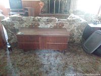 Front view of a wooden cedar chest with insert, showing the full chest against a floral patterned sofa on carpeted floor.