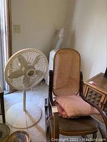 Photo showing the large cane backed rocking chair with pink seat cushion next to the white Lasko standing fan.