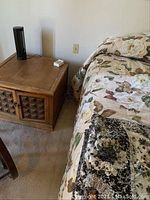 Wooden end table with square paneled doors, brass handles, on carpet next to king bed with floral bedding.