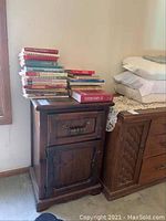 Dark wood cabinet with one drawer (missing handle) and one door, with a stack of over 25 books on top, mostly cookbooks and hardcovers, arranged in two piles with some visible titles.