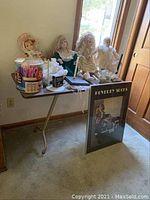 Four large vintage dolls seated on a folding table near window, with framed wall art and craft edgers nearby.