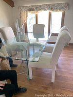 Full view of glass dining table with four white upholstered chairs around it in a bright room with hardwood floors.
