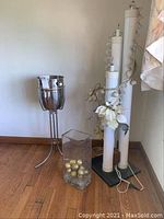 Full view of standing metal ice bucket, tall glass vase with gold ornaments, and three electric wedding candles decorated with string lights and white artificial flowers.