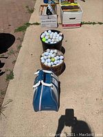 Two large containers filled with golf balls and a blue bowling ball bag on pavement.