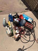 Overview photo of assorted auto supplies laid on a paved surface showing battery charger, fluid containers, funnels, cables, and traction cable box.