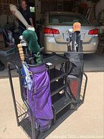 Two golf bags with clubs, one purple and one black TaylorMade, placed in a black metal stand with storage shelves, in a garage setting.