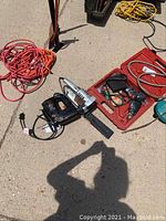Photo showing a reciprocating saw, cordless drill, small hand sander placed on ground next to coiled extension cords and a red toolbox.