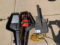 Overview of Homelite chainsaw in black case next to metal shop light with yellow cord on pavement