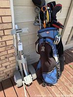 Blue and brown golf bag filled with multiple golf clubs including some with head covers, displayed next to the folded Bag Boy Lite hand cart on a wooden deck against a wall.
