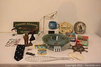 Full view of collection on table including pottery bowl signed 'Vicki', wooden plaques, metal balancing fisherman, Tommy Bahama mug, puzzles, tie, and trivet shaped like ship's wheel.