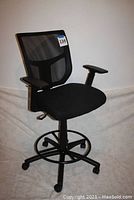 Side angle showing the black fabric seat, mesh backrest, adjustable armrests, five caster wheels, and footrest ring.