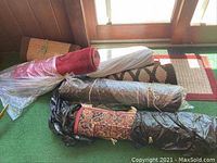 Photo showing multiple rolled rugs tied and wrapped, placed on green carpet near wooden windows.