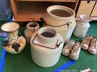 Overview of three ceramic crocks including two larger ones with metal handles, one plain smaller crock, decorative vase with grape and leaf design, and two pairs of beige baby shoes.