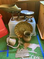 Wide view of lot with vintage metal scale on a raised surface, large metal pitcher in front, ornate metal plate behind, metal bell on the left, and Frog Crossing metal sign with attached cow silhouette at bottom front.