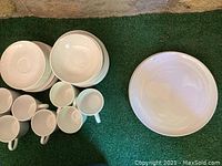 Set of white dishes including bowls, cups, and plates stacked on green surface, showing different shapes and sizes.