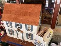 Front and side views of wooden dollhouse with shingled roof and blue window shutters, detailing construction and exterior features