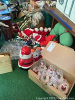 Overview of a variety of Christmas decor items on green surface including snow globes in box, ceramic Santa jar, glass tray, tin canister, wooden figurines, Santa hats and fabric wreath