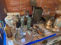 Photo showing assorted items including pewter pitcher, silver plated coffee pot, silver plated bucket, bottles and trays arranged on table.