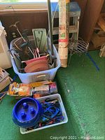 Binoculars case, blue dog bowl, leashes, hand warmers, various garden tools and stakes inside plastic containers on green carpeted floor near window.