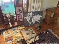 Photo showing an assortment of framed art pieces on the floor, including a world map and various cat themed prints, along with multiple wooden cat figurines on the floor and a piece of wooden furniture with heart cutout on drawer.