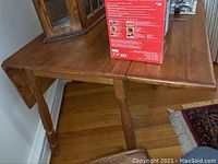 Top view of the wooden drop leaf table showing the surface and one foldable leaf with visible wear and scratches. A box on top is excluded from the sale.