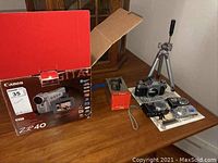 Lot overview showing Canon ZR40 camcorder box, Mico Vue device in orange box, Canon AE1 camera, tripod, Olympus Stylus 740 and one Panasonic Lumix camera on table