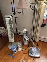 Photo showing two floor lamps on black wrought iron stands, white tower fan, oscillating fan, personal fan, Health O Meter scale and Lasko heater on wood floor.