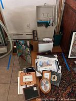 Photo showing assortment of picture frames, framed artwork and US quarter map, jewel cases, pliers, sunglasses, and wallets arrayed on floor and leaning against walls and furniture.