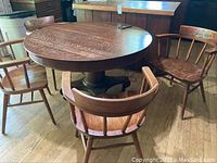 Photo showing round wooden dining table with pedestal base surrounded by four wooden chairs with spindled backs on wood floor.