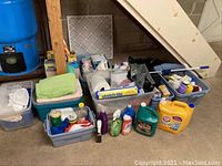 Plastic bins containing assorted cleaning products such as spray bottles, detergents, and cleaning liquids under stairs with large water barrel visible