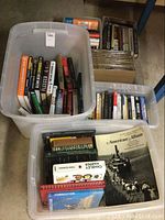 Two large clear plastic bins with multiple books inside, view from above showing diverse subjects including politics, history, and home improvement.