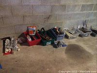 Wide photo showing multiple plastic bins containing tools, plumbing supplies, and various hardware items arranged on a concrete floor against a cinder block wall.