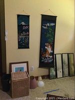 Photo showing wicker hamper, two Asian style hanging fabric tapestries, four framed art pieces leaning against wall, and three plastic lamp shades on floor.