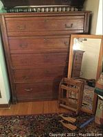 Photo showing wooden dresser with four drawers and framed mirror leaning against it, along with wooden mounted magazine rack on floor.